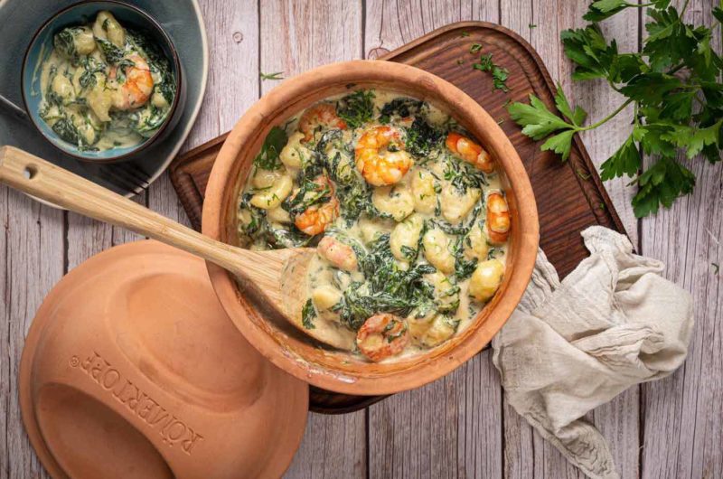 Gnocchi mit Garnelen und Spinat in cremiger Soße in einem Tontopf, daneben frische Petersilie und ein Leinentuch.