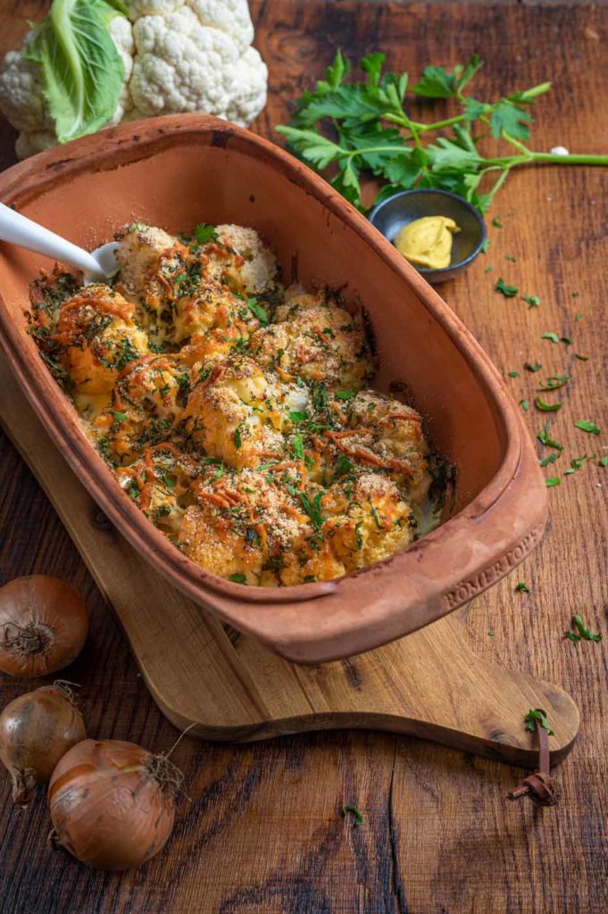Blumenkohlauflauf mit Käse und Kräutern, serviert in einer braunen Auflaufform auf einem Holzbrett, daneben Zwiebeln und Petersilie.