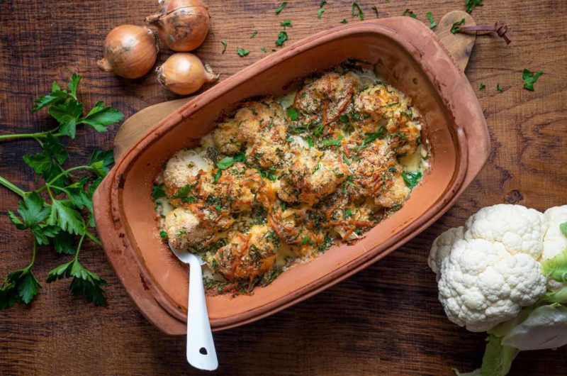 Blumenkohlauflauf mit Käse und Kräutern in einer Keramikform, daneben Zwiebeln, Petersilie und ein Blumenkohl auf Holzbrett.