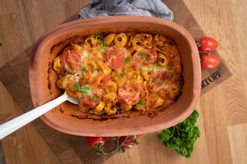 Keramikbecken mit überbackenen Tortellini, Tomaten und Käse, garniert mit Kräutern, auf Holzbrett serviert.