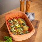 Keramikauflaufform mit gefüllten Zucchiniröllchen auf Tomatensauce, auf Holzbrett, daneben Petersilie und Salzstreuer.