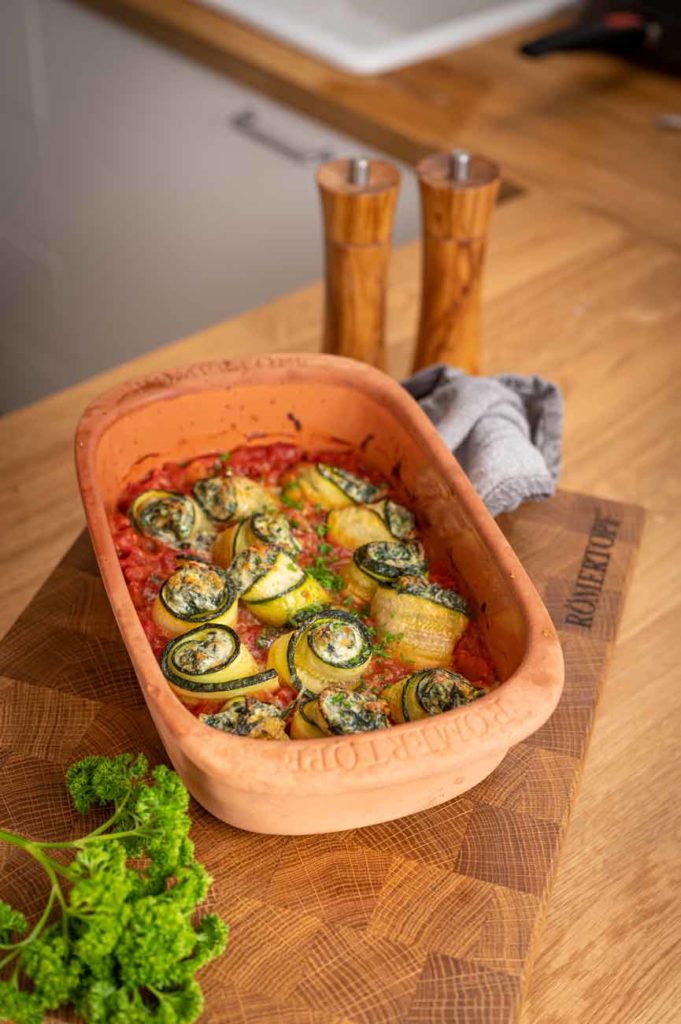 Keramikauflaufform mit gefüllten Zucchiniröllchen auf Tomatensauce, auf Holzbrett, daneben Petersilie und Salzstreuer.