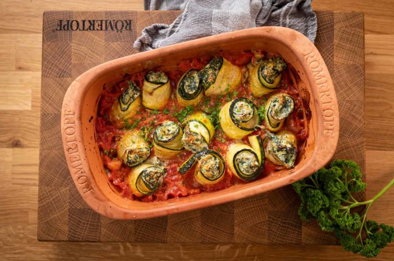 Auflaufform mit gefüllten Zucchini-Rollen, überbacken mit Käse, auf Tomatensauce. Petersilie daneben.
