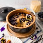 Goldbrauner Auflauf mit Blaubeeren und essbaren Blumen in einer Schüssel, Getränkekanne und Teller daneben.