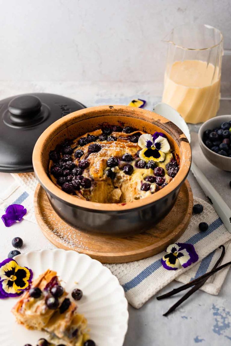 Goldbrauner Auflauf mit Blaubeeren und essbaren Blumen in einer Schüssel, Getränkekanne und Teller daneben.