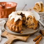 Ein angeschnittener Gugelhupf mit Zuckerguss auf einem Teller, daneben Zimtstangen und eine Gugelhupfform.