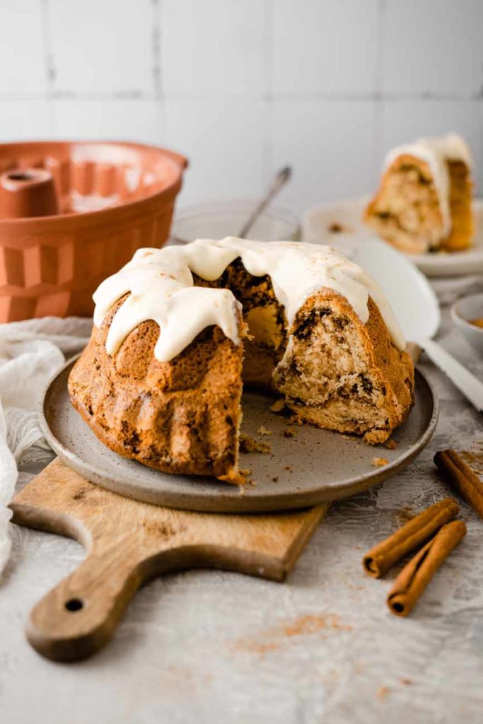 Ein angeschnittener Gugelhupf mit Zuckerguss auf einem Teller, daneben Zimtstangen und eine Gugelhupfform.