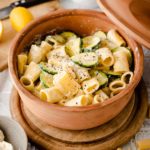 Rigatoni mit Zucchinis und Sahnesauce in einem braunen Keramiktopf, angerichtet auf einem Holzteller.