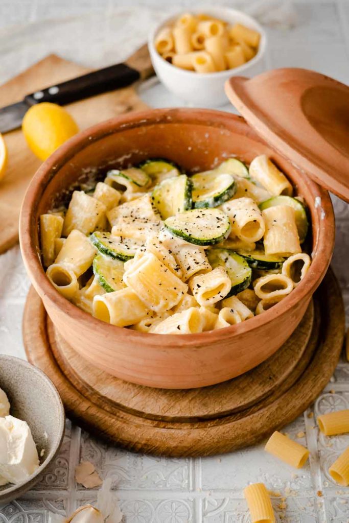 Rigatoni mit Zucchinis und Sahnesauce in einem braunen Keramiktopf, angerichtet auf einem Holzteller.