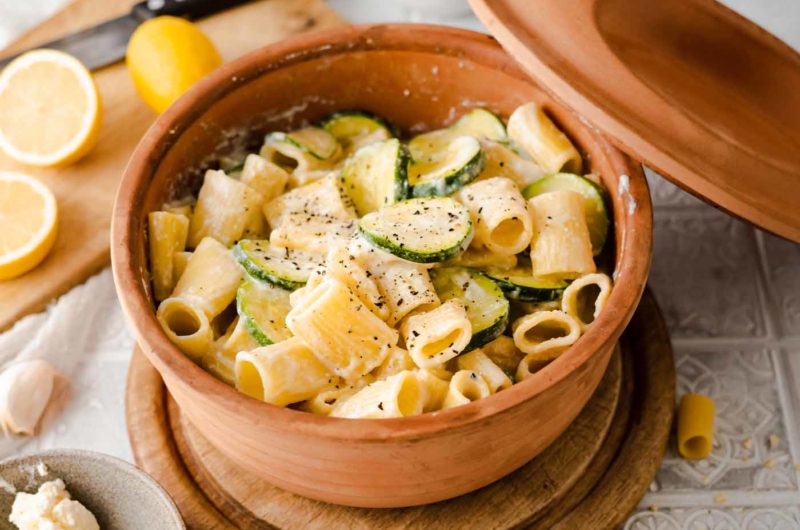 Rigatoni mit Zucchini und Sahnesoße in einem Tontopf, gewürzt mit Pfeffer. Im Hintergrund Zitrone und Knoblauch.
