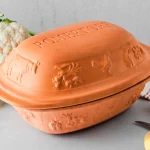 Ton-Römertopf mit Verzierungen, daneben Blumenkohl und Kartoffeln auf einem grauen Hintergrund.