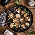 Gegrillte Auberginen auf schwarzem Teller, dekoriert mit Kräutern und Joghurtsauce, neben frischen Auberginen und Curry-Gewürz.