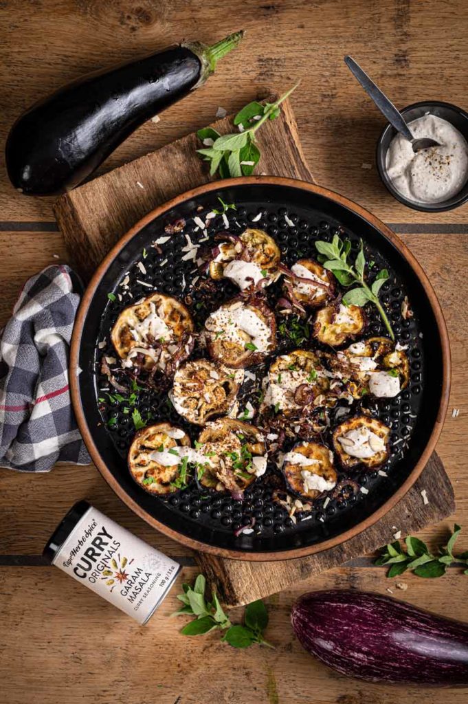 Gegrillte Auberginen auf schwarzem Teller, dekoriert mit Kräutern und Joghurtsauce, neben frischen Auberginen und Curry-Gewürz.