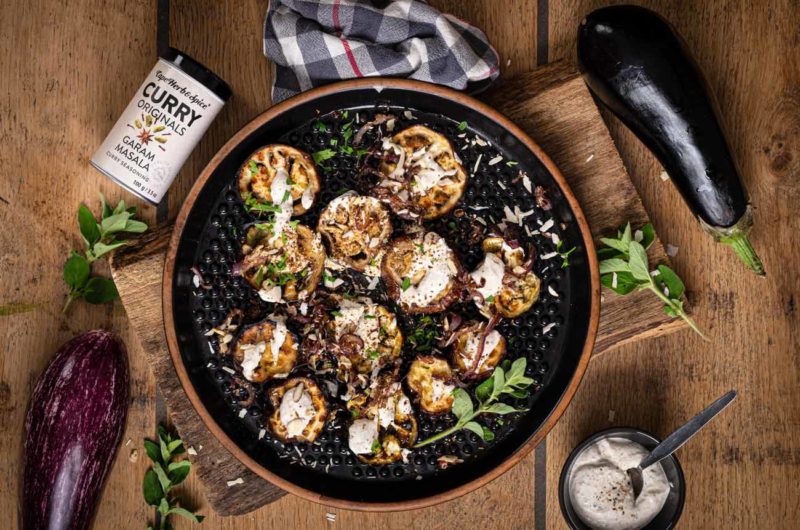 Gebratene Auberginenscheiben mit Sauce, Kräutern und Garam Masala in einer schwarzen Pfanne auf Holzbrett.