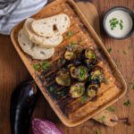 Gebratene Auberginenscheiben auf Teller mit Brot, Joghurt-Dip und frischen Kräutern, daneben zwei ganze Auberginen.