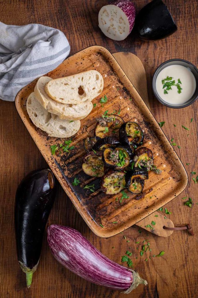 Gebratene Auberginenscheiben auf Teller mit Brot, Joghurt-Dip und frischen Kräutern, daneben zwei ganze Auberginen.