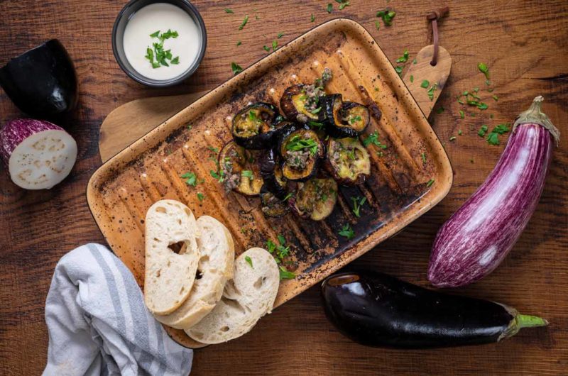 Gegrillte Auberginenscheiben auf einem Teller mit Brot und Dip auf einem Holztisch, dekoriert mit Petersilie.