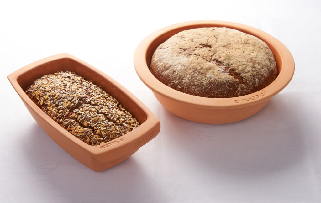 Zwei Brote in terrakottafarbenen Römertöpfen, eines mit Saaten, das andere mit Mehl bestäubt, auf weißem Tischtuch.