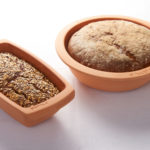 Zwei Brote in terrakottafarbenen Römertöpfen, eines mit Saaten, das andere mit Mehl bestäubt, auf weißem Tischtuch.