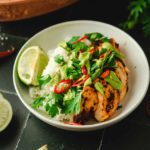 Schüssel mit gegrilltem Hähnchen, Reis, Avocado-Dip, Limettenscheibe, Chili und Petersilie auf dunklem Untergrund.