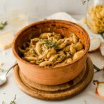 Bunter Keramiktopf mit Muschelpasta, garniert mit frischem Thymian, auf einem hölzernen Untersetzer.