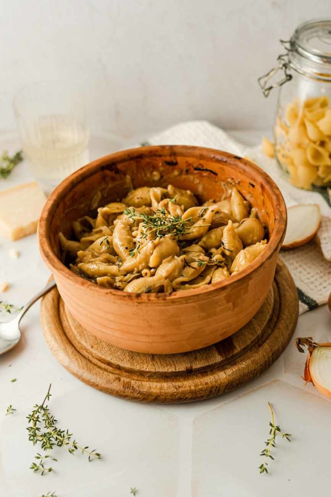 Bunter Keramiktopf mit Muschelpasta, garniert mit frischem Thymian, auf einem hölzernen Untersetzer.