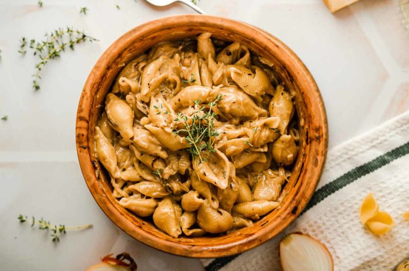 Schüssel mit Muschelnudeln in cremiger Soße, garniert mit frischem Thymian, auf weißem Untergrund mit Zwiebelstücken.
