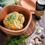 Gebackener Blumenkohl mit Tahini-Sauce in Tonform, umgeben von Knoblauch und frischen Kräutern auf Holzuntergrund.