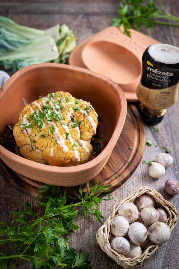 Gebackener Blumenkohl mit Tahini-Sauce in Tonform, umgeben von Knoblauch und frischen Kräutern auf Holzuntergrund.