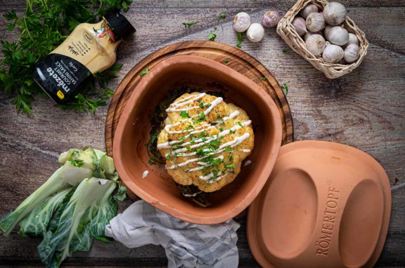 Römertopf mit gebackenem Blumenkohl, garniert mit Soße und Kräutern, umgeben von Senf, Knoblauch und frischem Grünzeug.