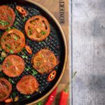 Gegrillte Tomatenscheiben mit Chili und Kräutern auf schwarzem Teller, daneben zwei frische rote Chilischoten.