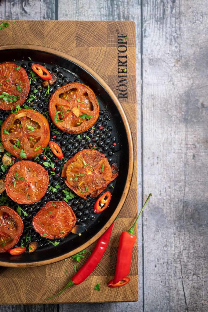Gegrillte Tomatenscheiben mit Chili und Kräutern auf schwarzem Teller, daneben zwei frische rote Chilischoten.