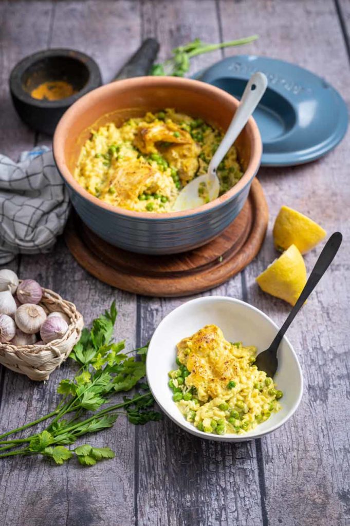 Schüssel mit cremigem Risotto, grünen Erbsen und gebratener Hähnchenbrust, daneben Knoblauch, Petersilie und Zitrone.