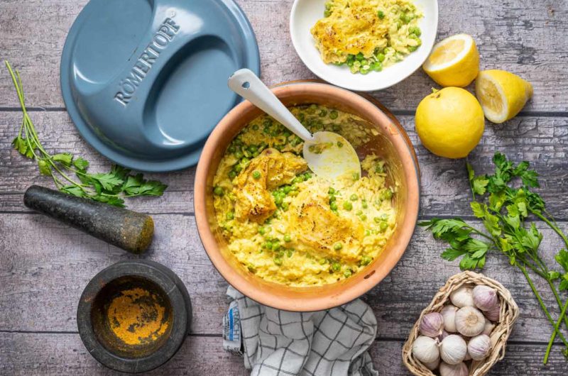 Ton-Topf mit Hähnchen und Erbsen-Risotto, umgeben von Petersilie, Knoblauch, Zitrone und einem Mörser auf Holztisch.
