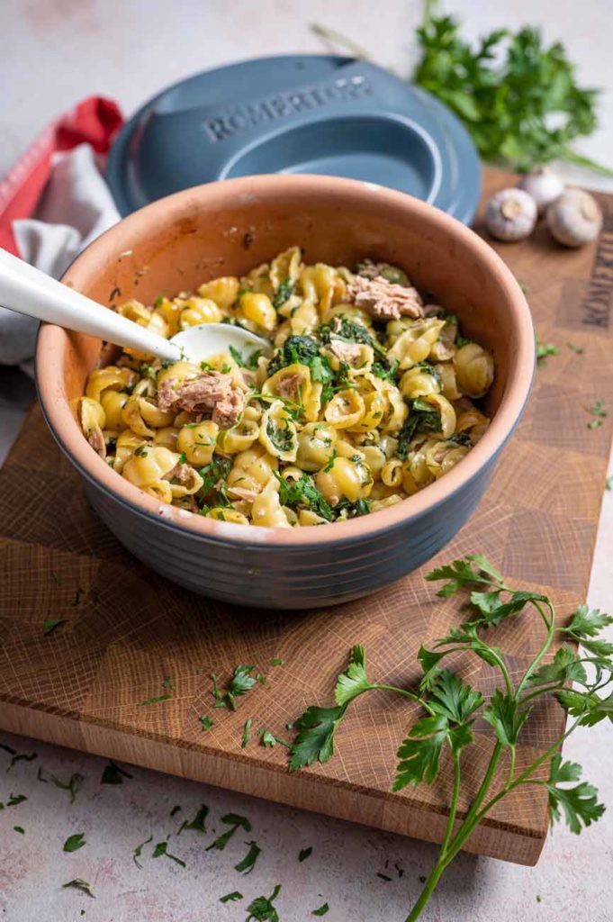 Schüssel mit Nudeln, Spinat, Thunfisch und Petersilie auf einem Holzbrett, daneben frische Kräuter.
