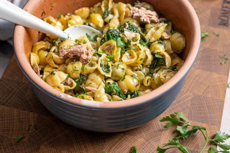 Schüssel mit Nudeln, Spinat, Thunfisch und Petersilie auf einem Holzbrett, daneben frische Kräuter.