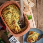 Gebratener Zucchini-Auflauf in einem Tontopf, daneben ein Teller mit einer Portion und Brot auf einem Holzbrett.