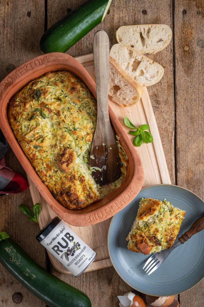 Gebratener Zucchini-Auflauf in einem Tontopf, daneben ein Teller mit einer Portion und Brot auf einem Holzbrett.
