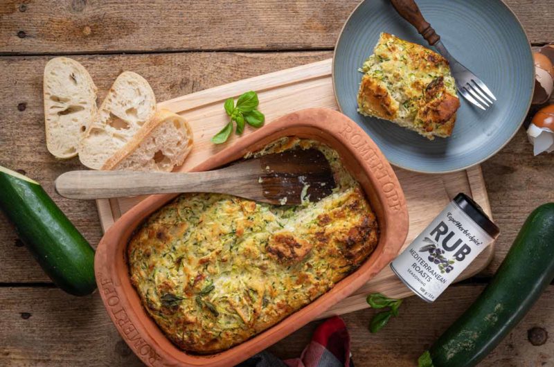 Auf Holztisch steht ein Gemüseauflauf in Römertopf, daneben Brot, Zucchini, Gewürz und ein Teller mit Auflauffstück.