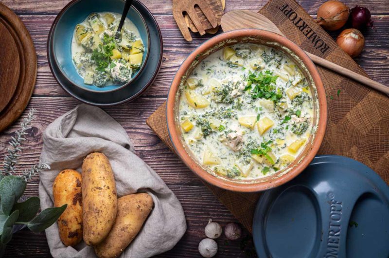 Eintopf mit Kartoffeln, Spinat und Sahne in einer Schüssel, daneben frische Kartoffeln und Zwiebeln auf Holztisch.