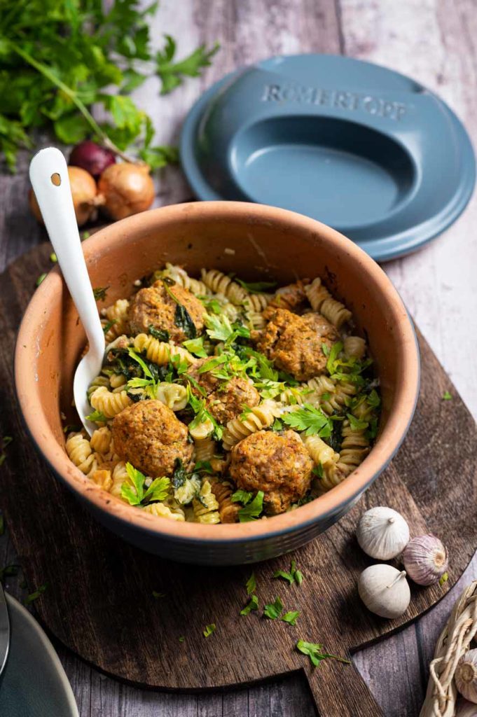 Keramikschale mit Nudeln, vegetarischen Bällchen und Kräutern auf Holztisch, daneben ein Römertopf-Deckel.