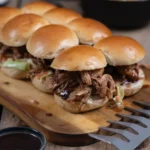 Mehrere Pulled-Pork-Burger auf einem Holzbrett, garniert mit Soße und Salat, daneben eine Metallbacke und ein Schälchen Soße.