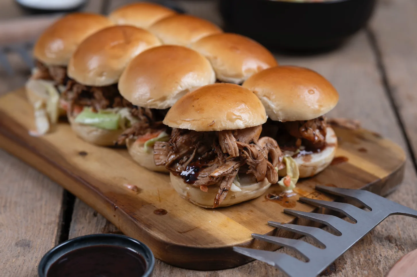 Mehrere Pulled-Pork-Burger auf einem Holzbrett, garniert mit Soße und Salat, daneben eine Metallbacke und ein Schälchen Soße.