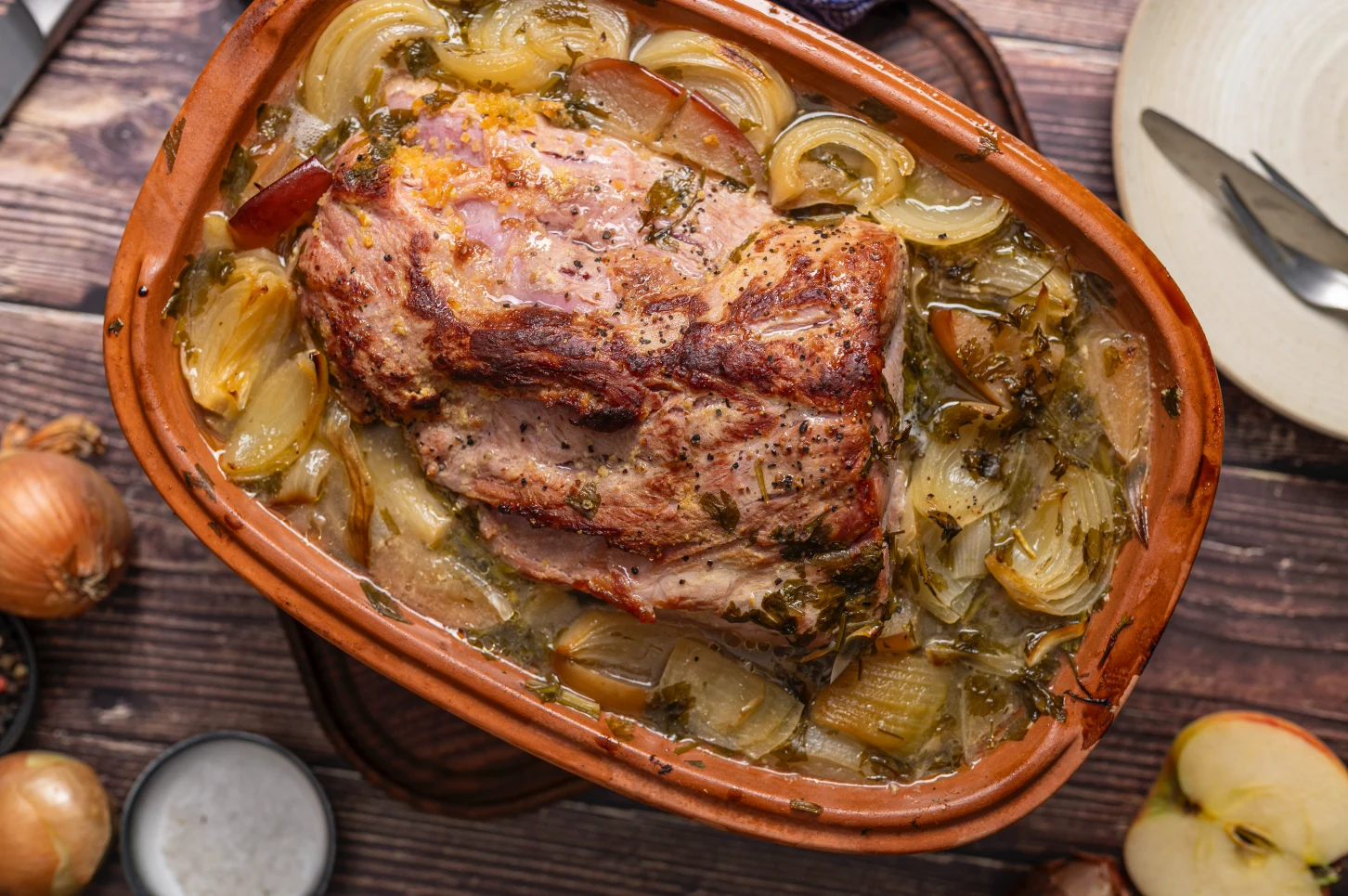 Braten mit Zwiebeln und Kräutern in Auflaufform auf Holzuntergrund, mit Zwiebeln und Äpfeln im Hintergrund.
