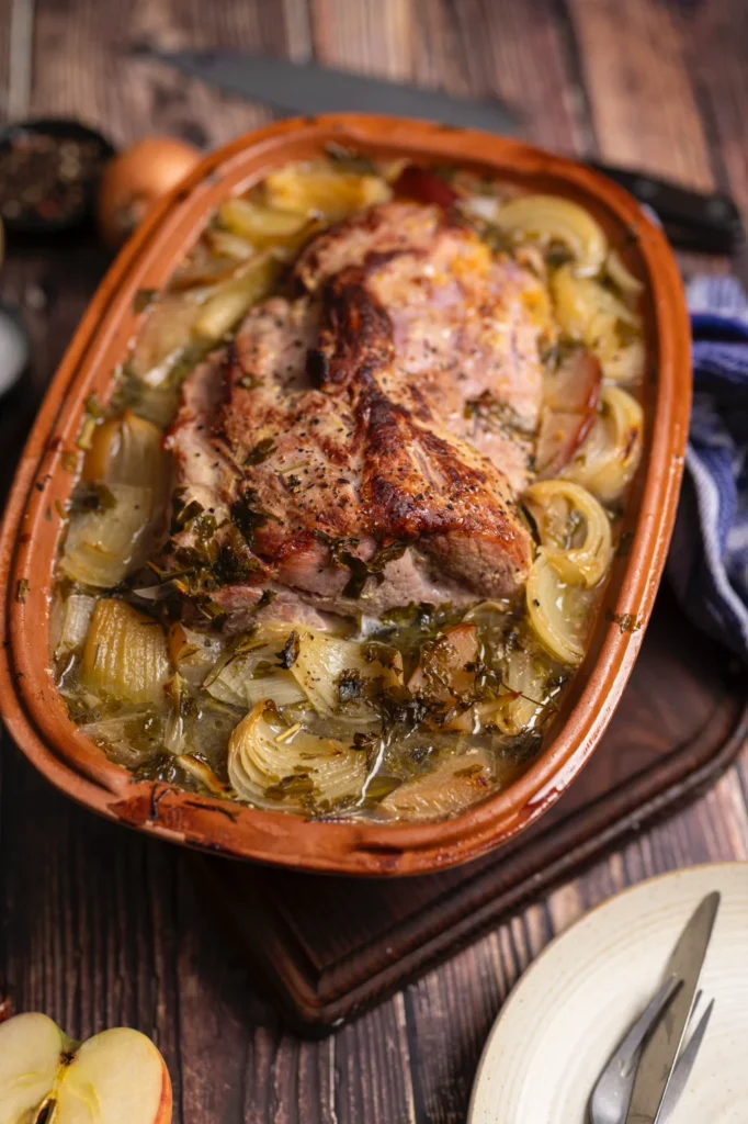 Bratenschüssel mit gebratenem Fleisch und Zwiebeln, umgeben von Kräutern, auf einem Holztisch serviert.