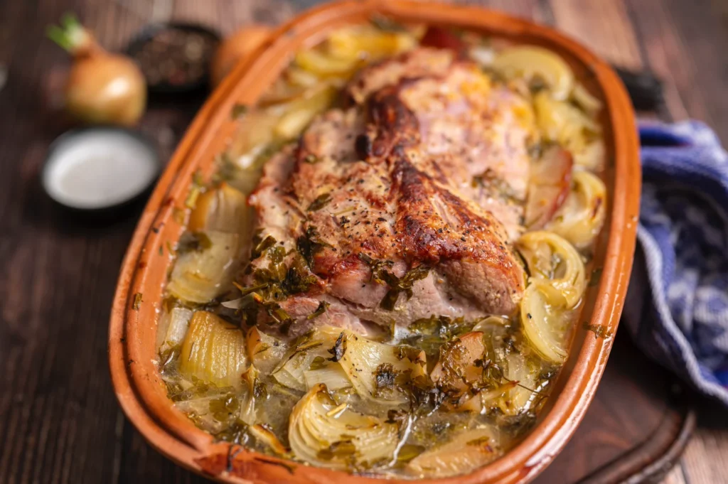 Bratenschale mit gebratenem Fleisch, Zwiebeln und Kräutern, auf einem Holztisch serviert. Hintergrund: Zwiebeln und Gewürze.