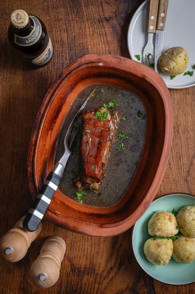 Gebackener Schweinebauch in einer Tonschale mit Kräutern, serviert neben Knödeln, Bierflasche und Besteck.