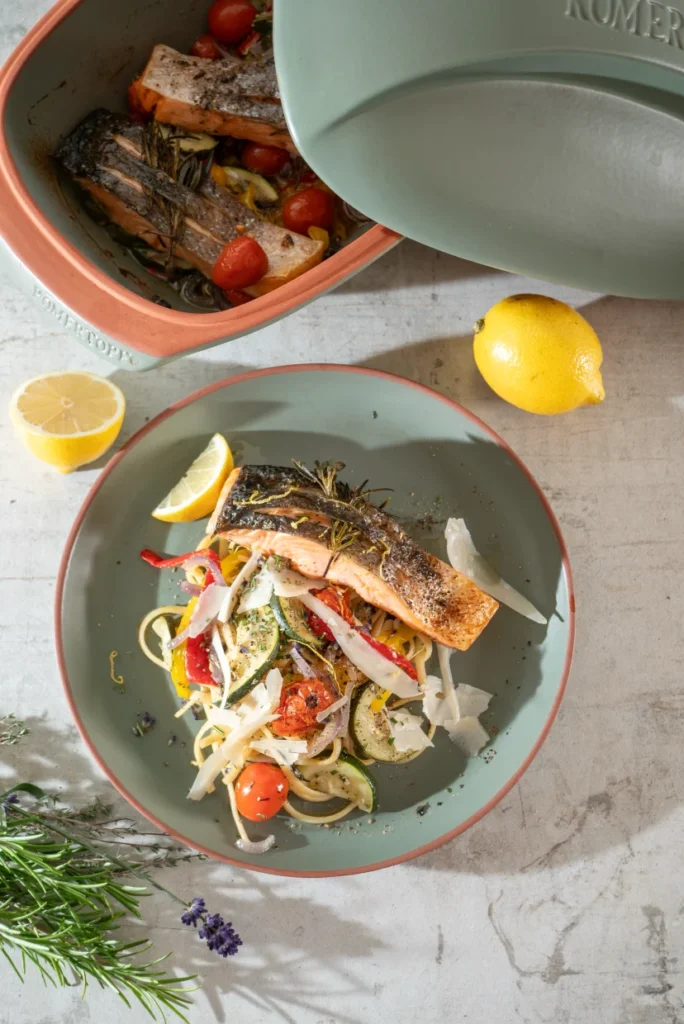 Gebratener Lachs auf Gemüse mit Rosmarin, serviert auf einem Teller mit Zitrone, im Hintergrund Römertopf und frische Kräuter.