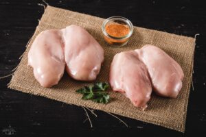 Rohe Hähnchenbrustfilets auf Sackleinen mit Petersilie und einer Schale Gewürz auf schwarzem Hintergrund.