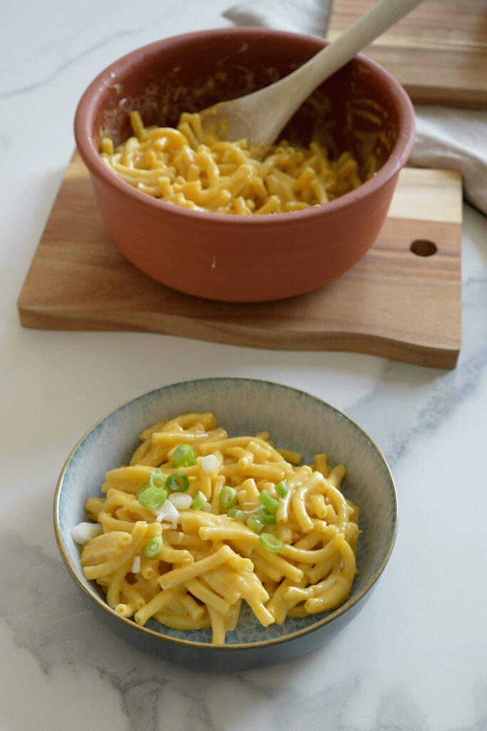 Zwei Schalen Mac and Cheese, eine im Vordergrund mit Frühlingszwiebeln garniert, eine auf einem Holzbrett im Hintergrund.